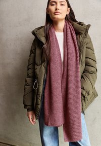 Bordeaux ribbel sjaal van textuurstof, gedragen over een witte top en groene pufferjack, gecombineerd met blauwe jeans.