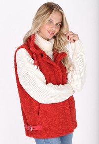 Rote, flauschige Weste mit hohem Kragen, Reißverschluss und Druckknopf-Details. Über einem weißen Strickpullover mit strukturierten Ärmeln getragen. Blaue Jeans.