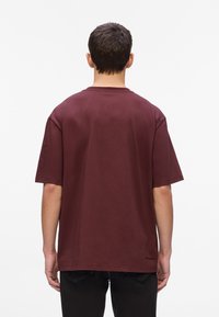 Burgundijas krāsas kokvilnas T-krekls, pārlieku liela izmēra, īsām piedurknēm, ar apaļu kakla izgriezumu, gluda tekstūra, mazs logotipa detaļas drēbju apakšā, valkāts kopā ar tumšām džinsām.