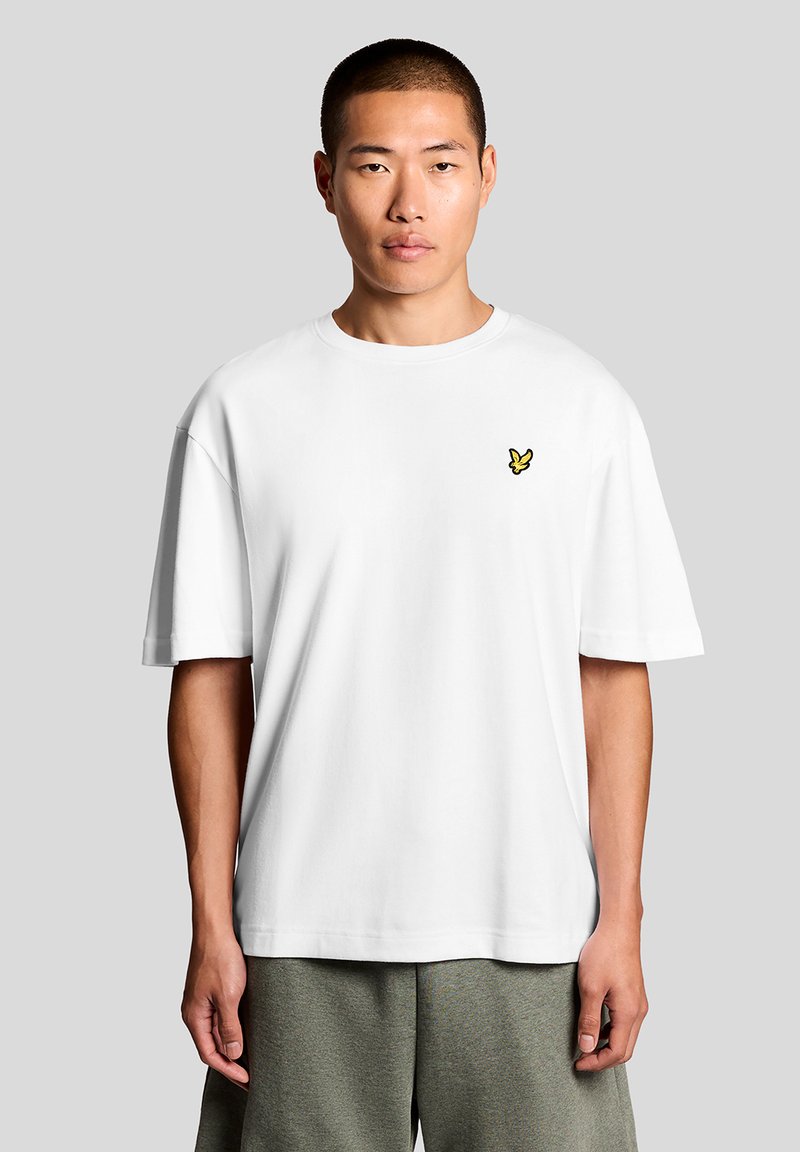 Homme portant un T-shirt blanc uni oversized avec un petit logo d'oiseau jaune sur le côté gauche de la poitrine, associé à un short gris ample.