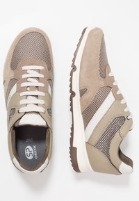 Beżowe buty sportowe z siateczkową cholewką i zamszowymi dodatkami. Białe paski po bokach, wyściełany kołnierz oraz gumowa podeszwa z teksturowanym bieżnikiem.
