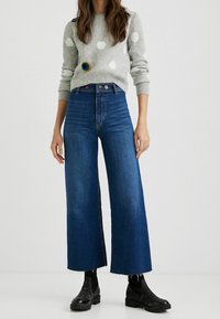 Pull gris à pois avec des accents brodés, associé à un jean en denim bleu à jambes larges, avec des ourlets effilochés et des bottines noires.