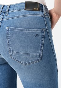 Blauwe denim jeans hebben een aansluitend ontwerp met een donker leren logo patch met de tekst "M.O.D." op de achterband en subtiele stiksels op de zakken.