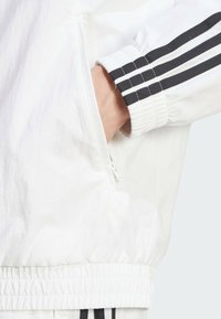 Vit zip-jacka med svarta tre-stripes accenter på ärmslutet. Har elastiska ärmslut och en avslappnad passform. Slät, lättviktsmaterial.
