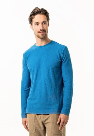 Man with short wavy hair wearing a blue long-sleeve shirt and beige pants, looking slightly to the side against a white background.