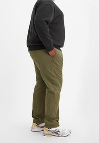 Olivengrüne Hose mit geradem Schnitt und seitlichen Taschen, kombiniert mit einem schwarzen Sweatshirt und weißen Sneakers mit gelben Akzenten.