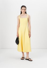 Vestido midi de lino amarillo con escote cuadrado y tirantes anchos, combinado con un bolso negro y chanclas negras. Textura suave, silueta en A.