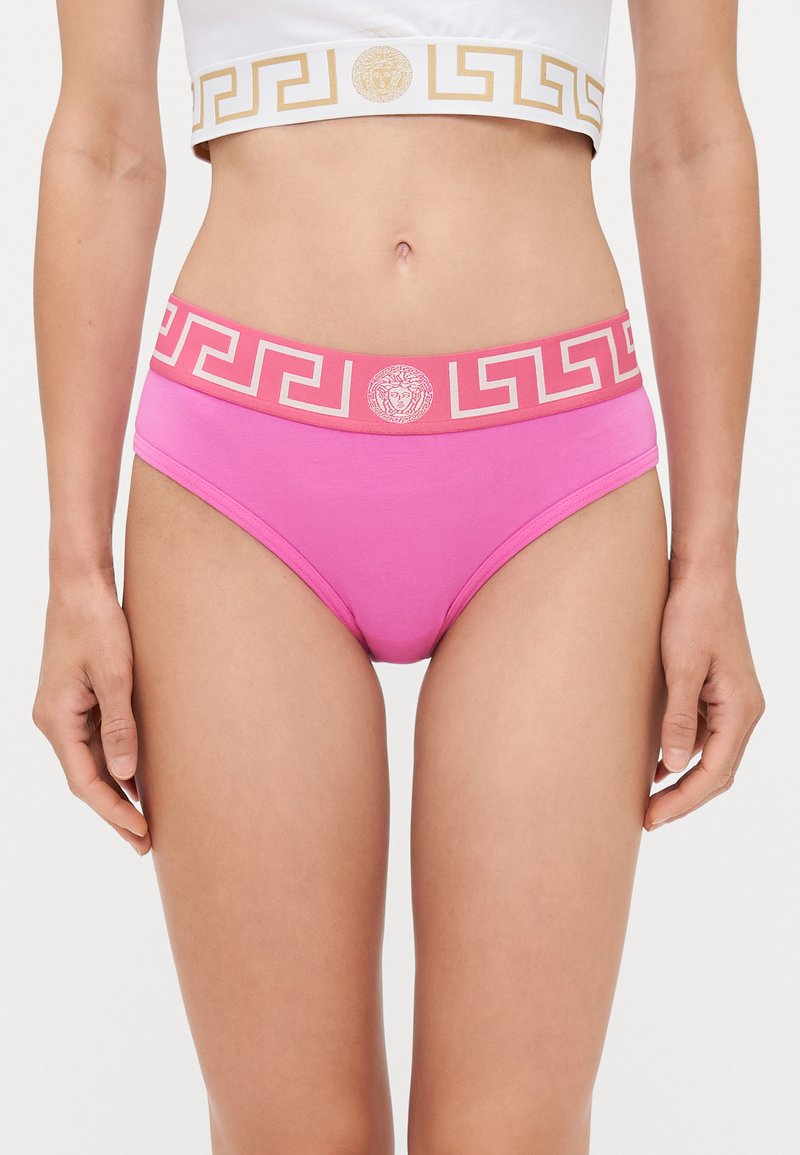 Rozā kokvilnas apakšveļa ar plašu jostu, uz kuras ir ģeometrisks dizains un logo. Josta ir papildināta ar kontrastējošu balto crop top.