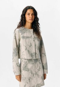 Veste courte en tissu beige avec des motifs floraux et d'oiseaux verts, dotée d'un encolure ronde et d'une fermeture à boutons. L'ensemble comprend une jupe assortie.