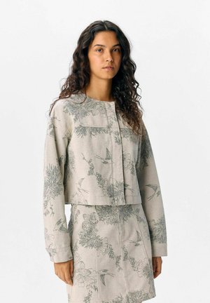 Veste courte en tissu beige avec des motifs floraux et d'oiseaux verts, dotée d'un encolure ronde et d'une fermeture à boutons. L'ensemble comprend une jupe assortie.