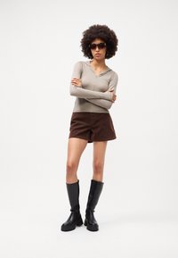 Langærmet taupe top med lynlås, brune ruskinds shorts og sorte knæhøje støvler. Accessoires inkluderer mørke solbriller og hoops-øreringe.