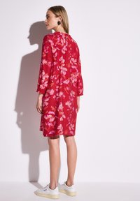 Rotes Blumenkleid mit dreiviertel Ärmeln, das mit pinken und orangenen Mustern verziert ist. Das Model trägt weiße Sneakers, die einen lässigen Sitz und eine Länge über dem Knie zeigen.