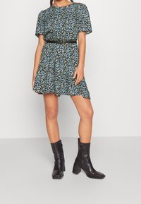 Vestido negro de flores de manga corta con patrones azules y amarillos, cintura ajustada con un cinturón negro, combinado con botas negras de tacón medio.