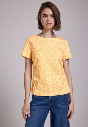 Chemise jaune à manches courtes avec un petit motif floral blanc. Tissu doux, encolure ronde et coupe légèrement décontractée. Associée à un jean bleu.