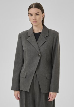Frau in einem grauen maßgeschneiderten Blazer mit breiten Revers, geknöpften Vorderteil und passenden Weitbeinhosen vor einem einfarbigen grauen Hintergrund.
