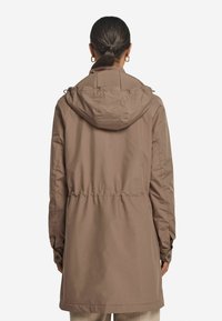 Brun, vattentålig parka med cinchad midja, huva och tryckknappsmanschetter. Har en smidig textur och en något oversize passform.