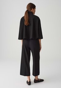 Black cropped cape-style jacket with a high neck, paired with wide-leg black trousers and black mesh slip-on shoes. Smooth texture, minimal design.