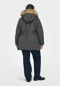 Pelēka puffer jaka ar mākslīgo kažokādu apdari uz kapuces, regulējama sasaitē josta, sānu pogas un redzamie šuvju sīkumi. Valkāta virs tumšajām džinsām.
