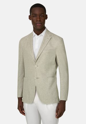 Homme portant un blazer texturé beige clair par-dessus une chemise blanche à col et un pantalon blanc, debout devant un fond uni clair.