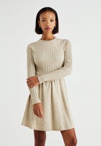 Robe en tricot beige avec une texture côtelée, des manches longues et un buste ajusté qui se transforme en une jupe évasée. Finition en tissu lisse.