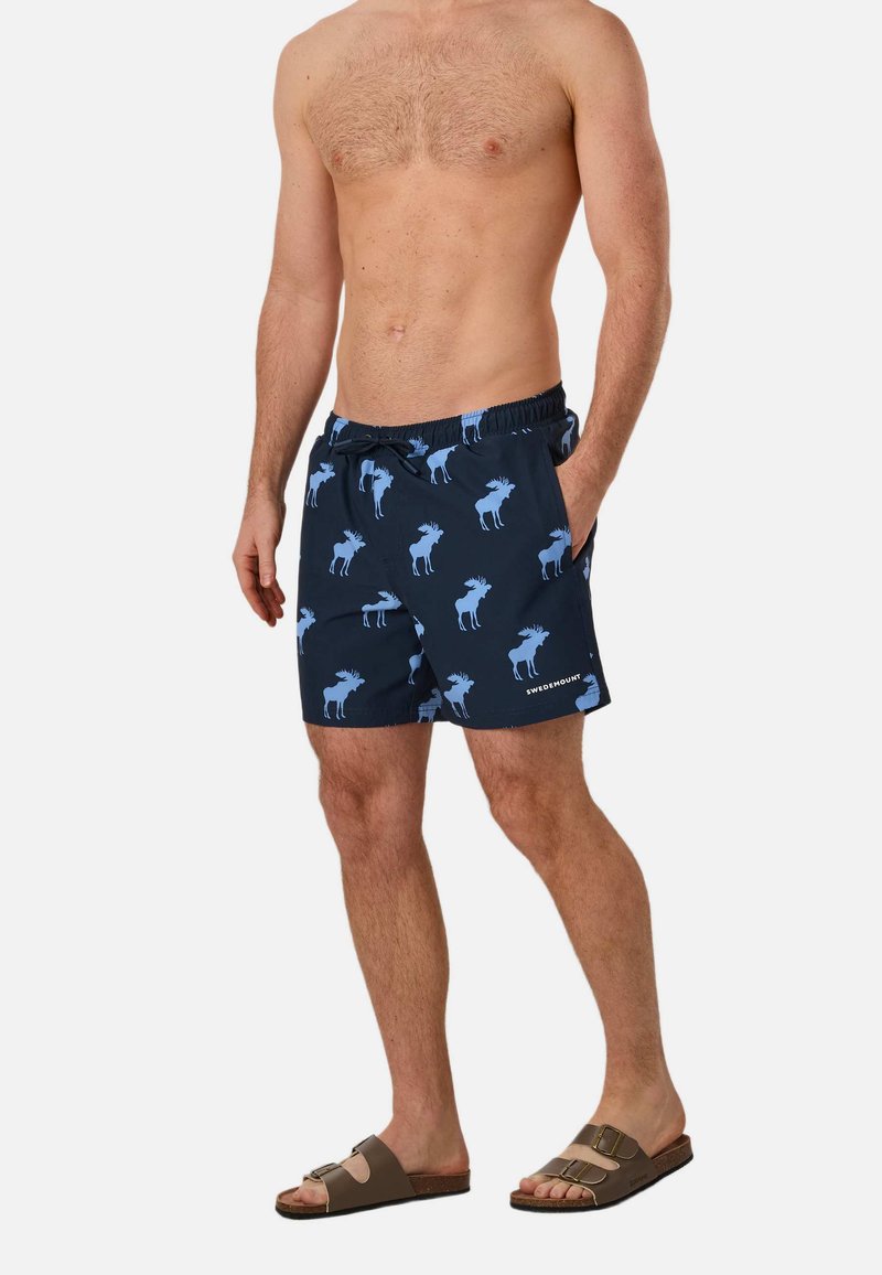 Homme torse nu portant un short de bain bleu marine avec des motifs élan bleu clair et des sandales marron à double bride, debout avec une main dans la poche.