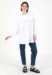Weiße Bluse mit langen Ärmeln und Rüschendetails an den Schultern, geraffter Vorderseite, kombiniert mit blauen Jeans und marineblauen Sandalen.