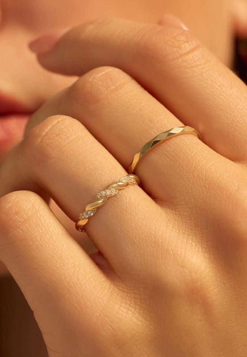 Deux bagues sur une main : l'une est un anneau en or torsadé orné de petits diamants, l'autre est un anneau en or plat martelé avec un design géométrique.