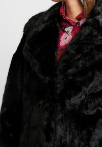 Černý kabát z umělé kožešiny s širokým límcem ve tvaru V, s jemnou texturou a hladkým leskem, zobrazený částečně přes top s květinovým vzorem.