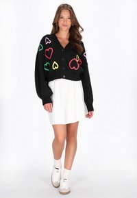 Svart kort cardigan med flerfärgade hjärtan, med V-ringning och knappstängning, bärs över en vit plisserad klänning och sneakers.