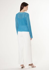 Leichter, offener Strickpullover in Türkis mit langen Ärmeln, rundem Ausschnitt und weißen Akzenten, kombiniert mit weiten weißen Hosen und Absatzsandalen.