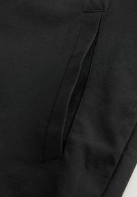 Los pantalones de tela negra presentan un bolsillo lateral con un cierre de cremallera oculto. El material parece suave con un ligero brillo, y se observan mínimos detalles de costura.