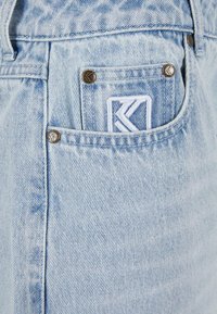 Heledad sinine denimist valmistatud teksapüksid, kontrastõmblusega, ees oleva väikese tasku ja valge broderitud logoga. Näha on metallist tarvikute aktsente.