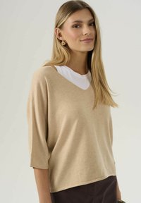Žena s dlouhými blond vlasy, oblečená v béžovém svetrě s výstřihem do V přes bílou halenku a tmavé kalhoty, s mírným úsměvem na jednoduchém pozadí.