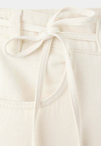 Pantalon en jean blanc avec un cordon noué à la taille et des coutures visibles sur les poches avant.