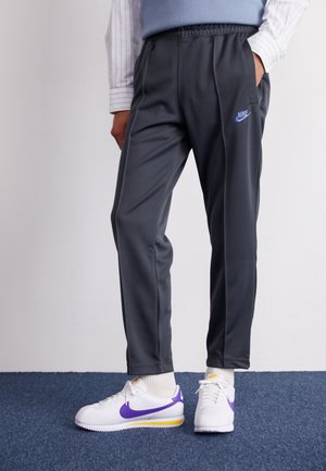 Mand iført mørkegrå Nike træningsbukser og hvide Nike sneakers med lilla swooshes står på et blåt tæppe.