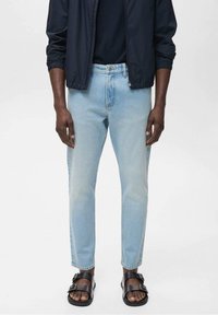 Vaaleansiniset farkut suoralla lahkeella, haalistuneella kuvioinnilla ja viiden taskun tyylillä, yhdistettyinä mustiin sandaaleihin ja tummansiniseen takkiin.