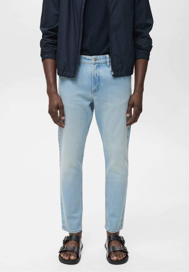 Vaaleansiniset farkut suoralla lahkeella, haalistuneella kuvioinnilla ja viiden taskun tyylillä, yhdistettyinä mustiin sandaaleihin ja tummansiniseen takkiin.