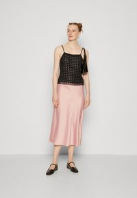 Svart ermeløs topp med hvite prikker, kombinert med en lys rosa midi skjørt. Modellen har på seg svarte leiligheter. Enkelt, elegant design med glatte teksturer.