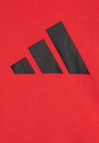 Tejido de algodón rojo con un gran y audaz diseño de logo negro de tres franjas. La textura es suave y ligeramente reflectante.