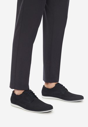 Czarne buty zamszowe i materiałowe z zaokrąglonym noskiem, sznurowane, z kontrastową białą podeszwą, zestawione z ciemnymi spodniami.