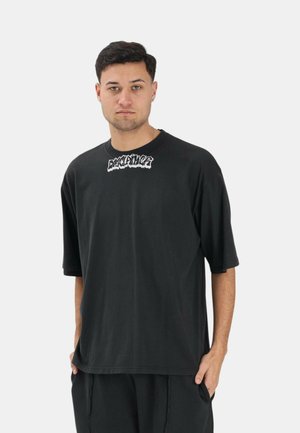 Uomo con capelli corti e scuri che indossa una maglietta nera oversize con scritta grafica bianca "EXCLAIMER" sul petto, in piedi davanti a uno sfondo chiaro uniforme.