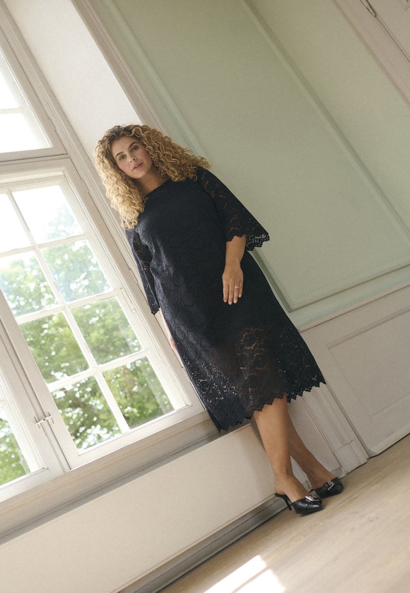 Robe en dentelle noire à manches trois-quarts et ourlet festonné. Portée avec des talons noirs brillants, devant un fond lumineux de fenêtre.
