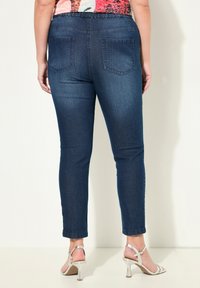 Úzké džíny v tmavě modrém denimu s zadními kapsami, s hladkým povrchem a přiléhavým střihem, doplněné o stříbrné sandály na vysokém podpatku.