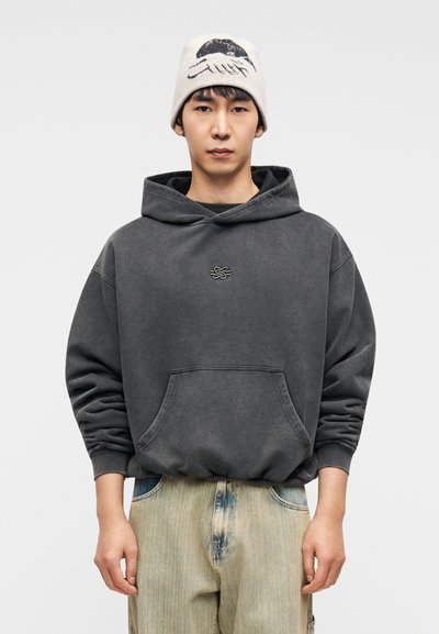 Σκούρα γκρι μεγάλο φούτερ με μπροστινή τσέπη και ριμπ μανσέτες, σε συνδυασμό με ανοιχτό μπλε τζιν παντελόνια με ελαφρώς φθαρμένο ύφος και εκρού σκουφάκι με μαύρο σχέδιο.