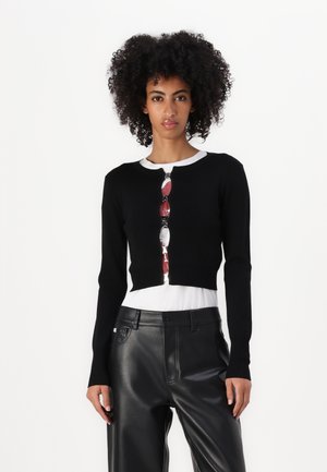 Cárdigan negro corto hecho de una tela suave, con cuello redondo y detalle de botones, superpuesto a una camisa blanca y combinado con pantalones negros.