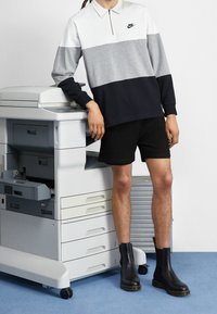 Nike långärmad polo med vita, grå och marinblå färgblock; svarta shorts; svarta stövlar; bredvid en multifunktionsskrivare.