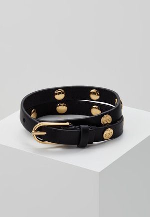 Ceinture en cuir noir enroulée sur une surface blanche avec une boucle dorée et des clous ronds dorés régulièrement espacés sur toute la longueur.