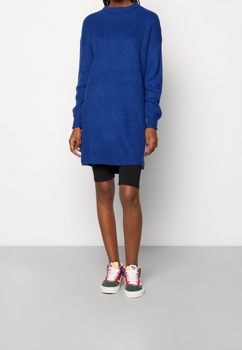 Personne portant un long pull en laine bleu, un short noir jusqu'aux genoux et des baskets colorées, debout contre un mur blanc uni.