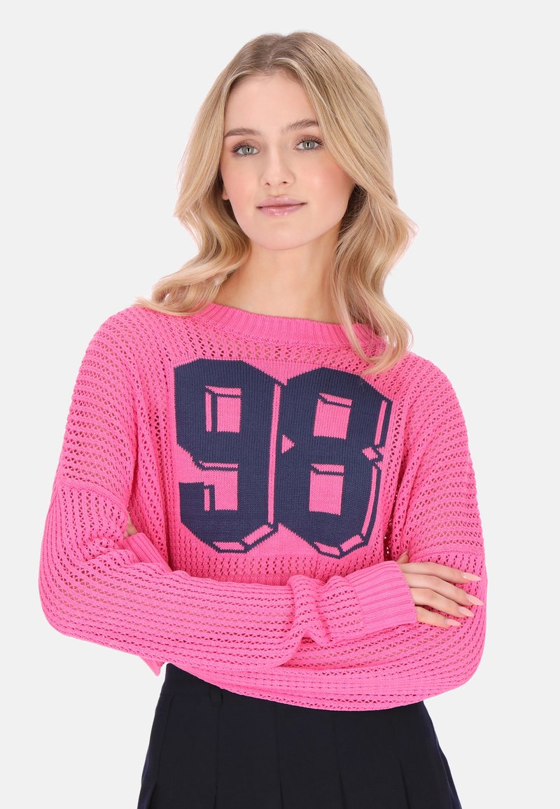 Fiatal nő szőke hajjal, rózsaszín kötött pulóvert visel, sötétkék "98" szám szerepel rajta, karjait keresztbe téve áll fehér háttér előtt.