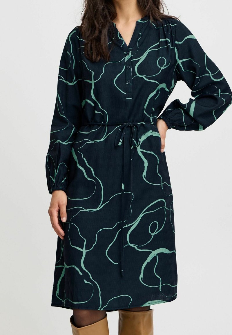 Femme portant une robe noire mi-longue avec des motifs de lignes abstraites vertes, manches longues, boutons au niveau de l'encolure et ceinture à la taille, associée à des bottes couleur beige.